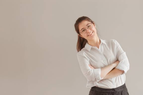 Junge Frau mit Brille steht lächelnd mit verschränkten Armen.